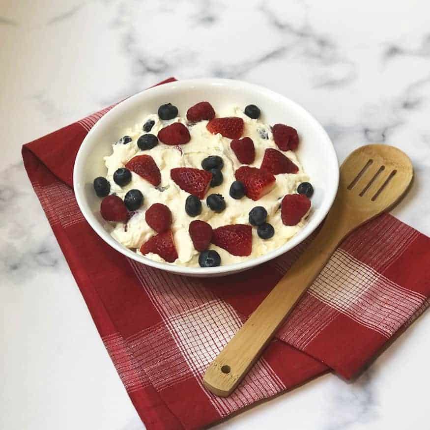 Triple Berry Cheesecake Fruit Salad in a white bowl with a wooden spoon and red linen under it. 