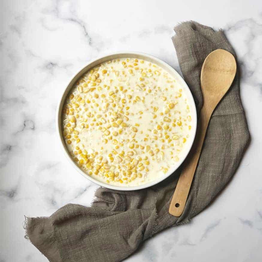 Rudy's BBQ Creamed Corn in a white dish with a gray linen and wooden spoon near it. 