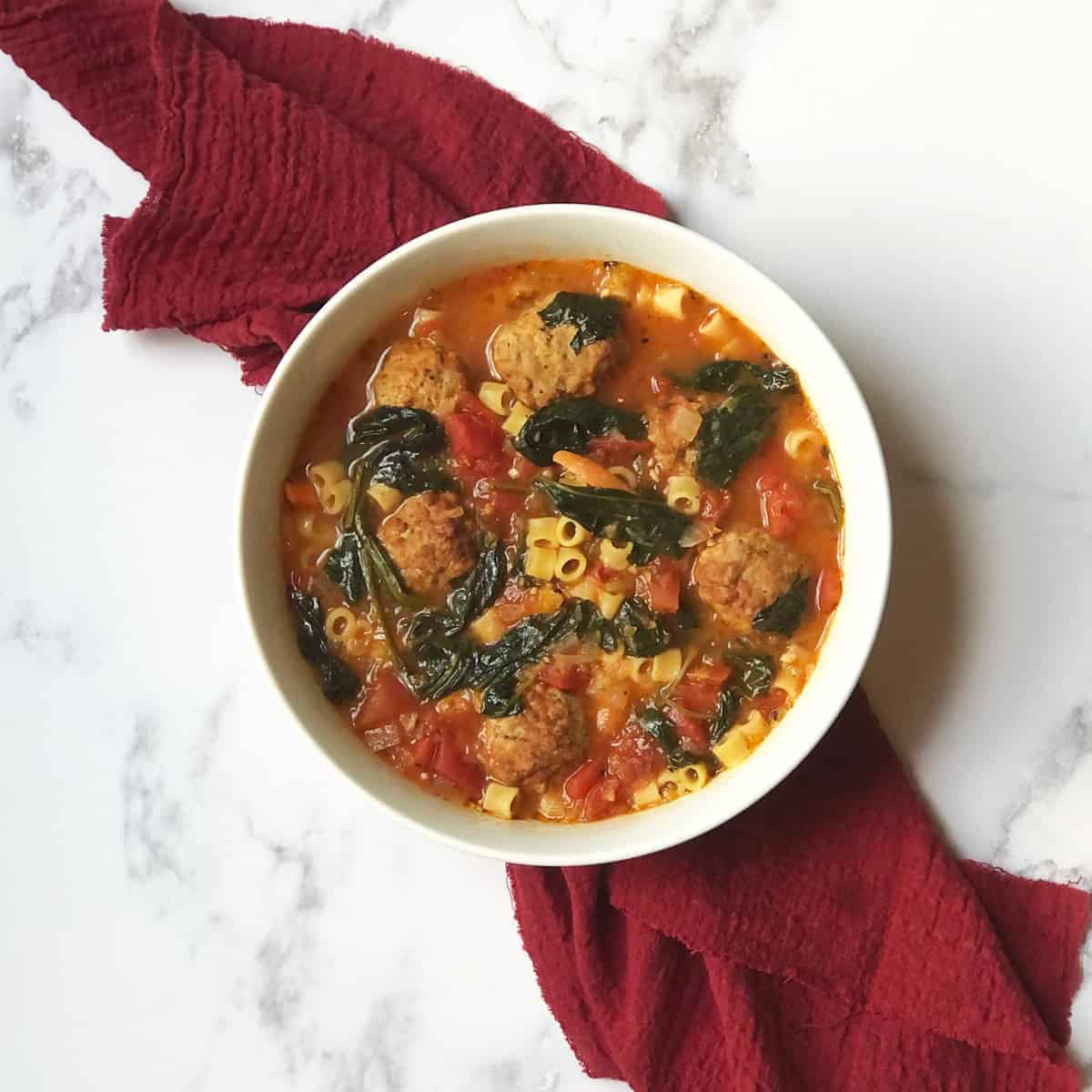 Lactation Soup (soup with spinach, meatballs, and pasta) in a white bowl on a red linen