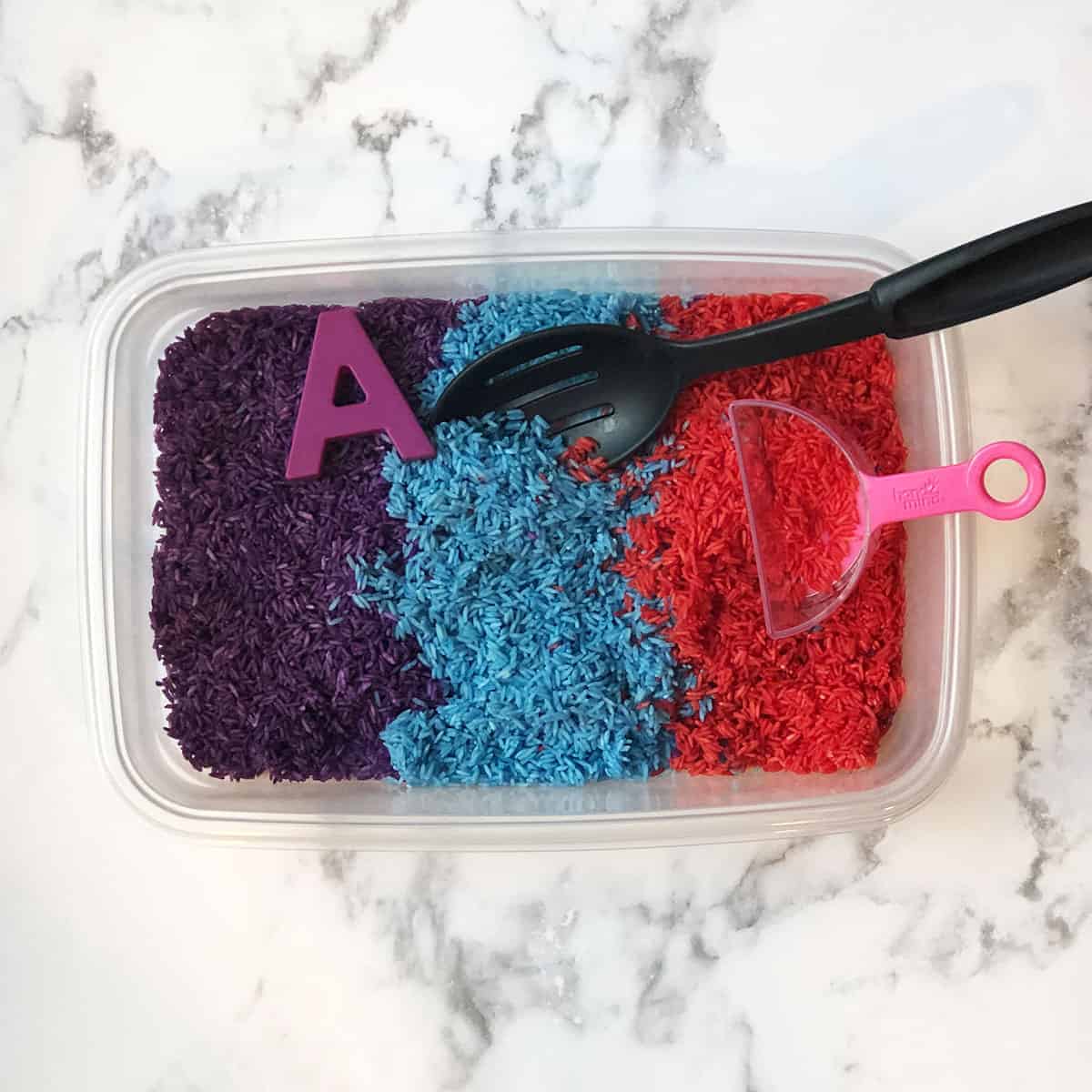 Sensory bin with purple, blue, and dark pink dyed rice with a spoon, letter, and measuring cup. 
