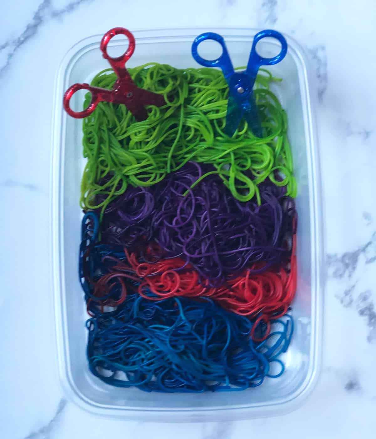 Spaghetti noodles that have been dyed green, purple, pink, and blue in a sensory bin with two pairs of safety scissors