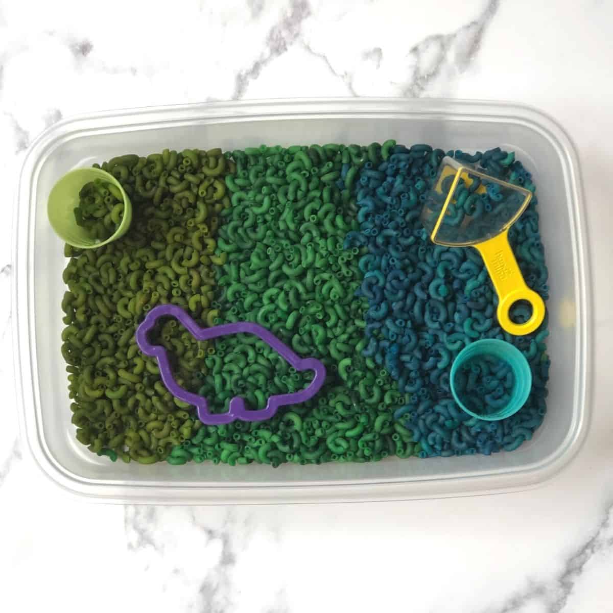 dyed macaroni noodles in a sensory bin with two cups, a measuring cup and a dinosaur cookie cutter